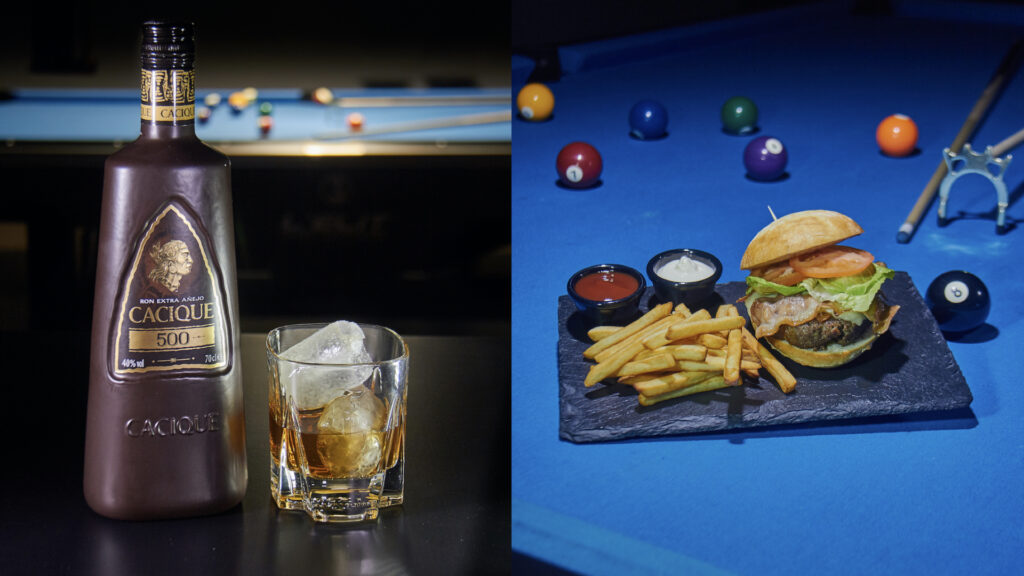 Botella de ron cacique con vaso lleno de ron y hielo al lado de una hamburguesa sobre una mesa de billar